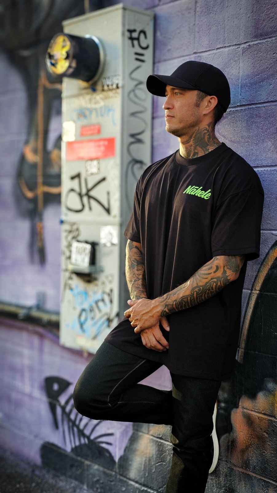 Concrete Bloom - Pulse of Kakaʻako Collection - Nāhele Clothing Co. - Nahele Co. - Nahele Clothing Co. - Man wearing a black t-shirt with green text and black pants leaning against a graffiti - covered wall.