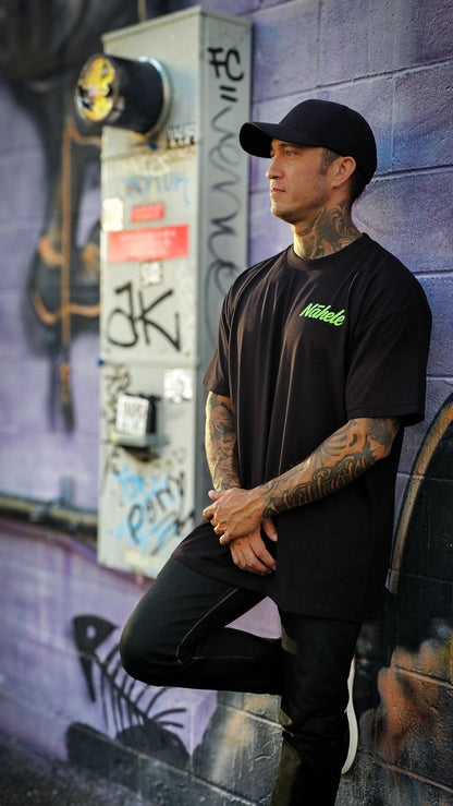 Concrete Bloom - Pulse of Kakaʻako Collection - Nāhele Clothing Co. - Nahele Co. - Nahele Clothing Co. - Man wearing a black t-shirt with green text and black pants leaning against a graffiti - covered wall.