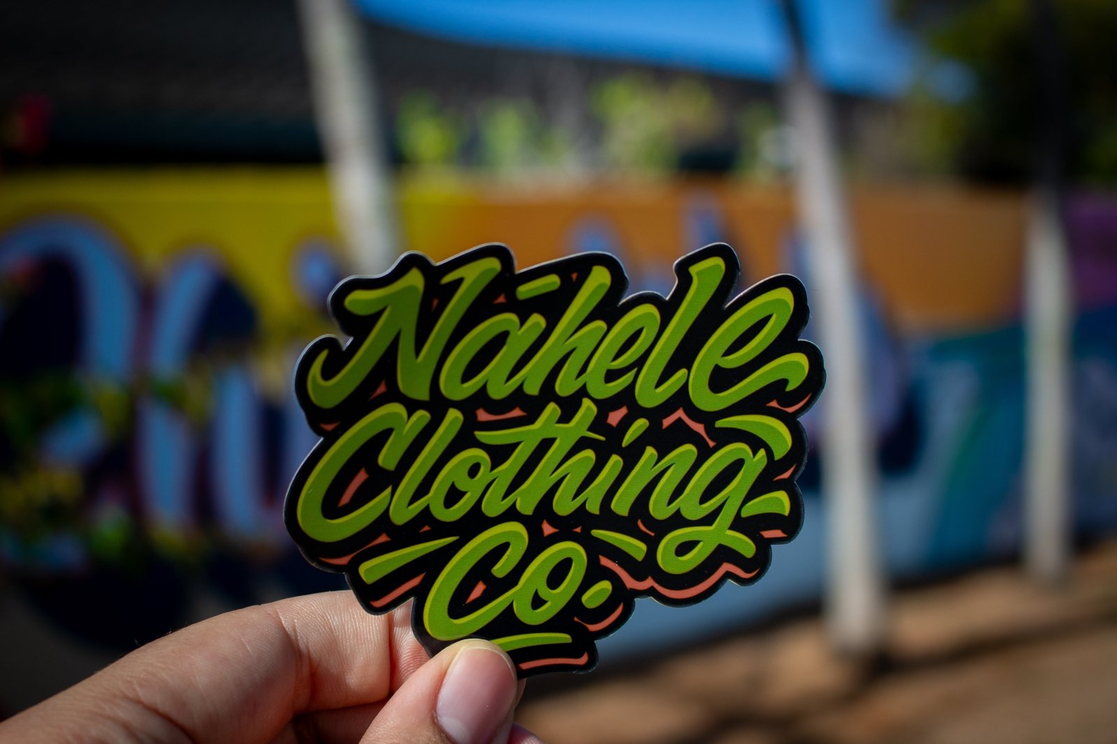 Nāhele Clothing Co. “Concrete Bloom” Sticker - Nāhele Clothing Co. - Nahele Co. - Nahele Clothing Co. - Sticker with 'Nahele Clothing Co.' text held in front of a colorful blurred grafitti style background